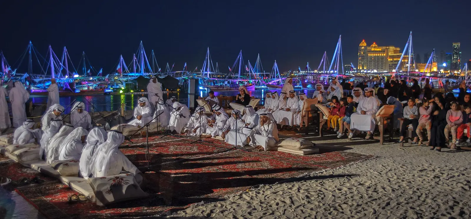 Performers seated on carpets at night on Katara beach with illuminated dhow masts behind
